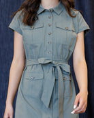 A woman wearing a sage green short-sleeve shirt dress with a matching tie belt, shown from the neck down against a dark blue textured background.