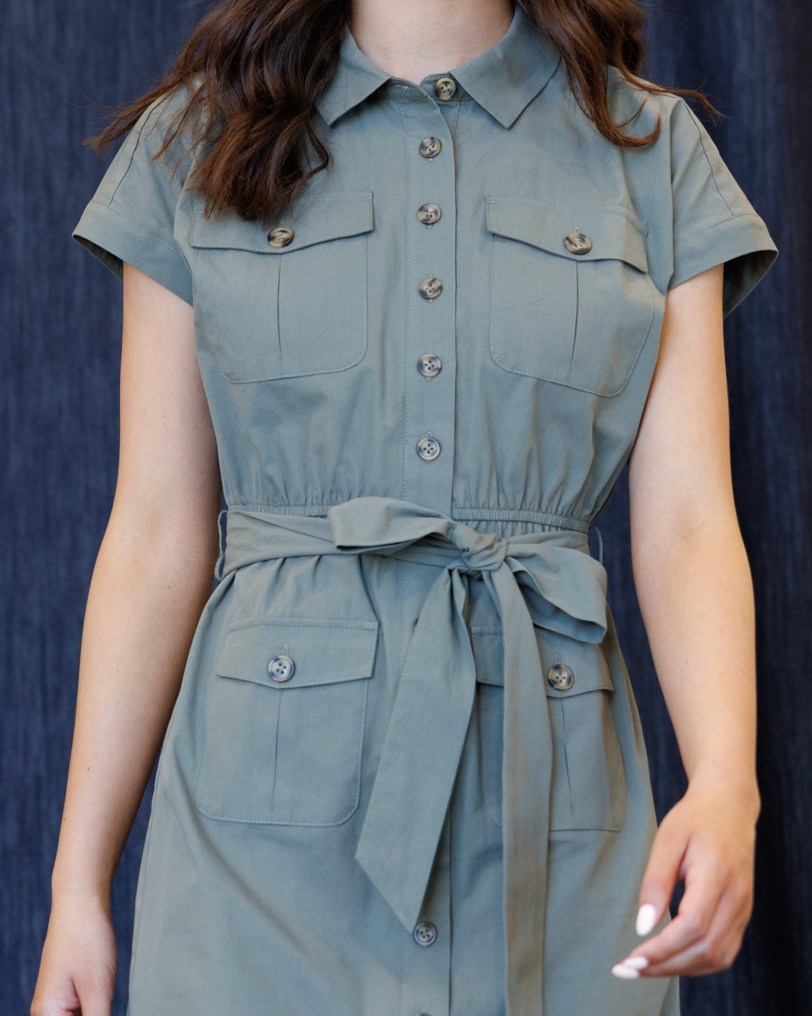 A woman wearing a sage green short-sleeve shirt dress with a matching tie belt, shown from the neck down against a dark blue textured background.