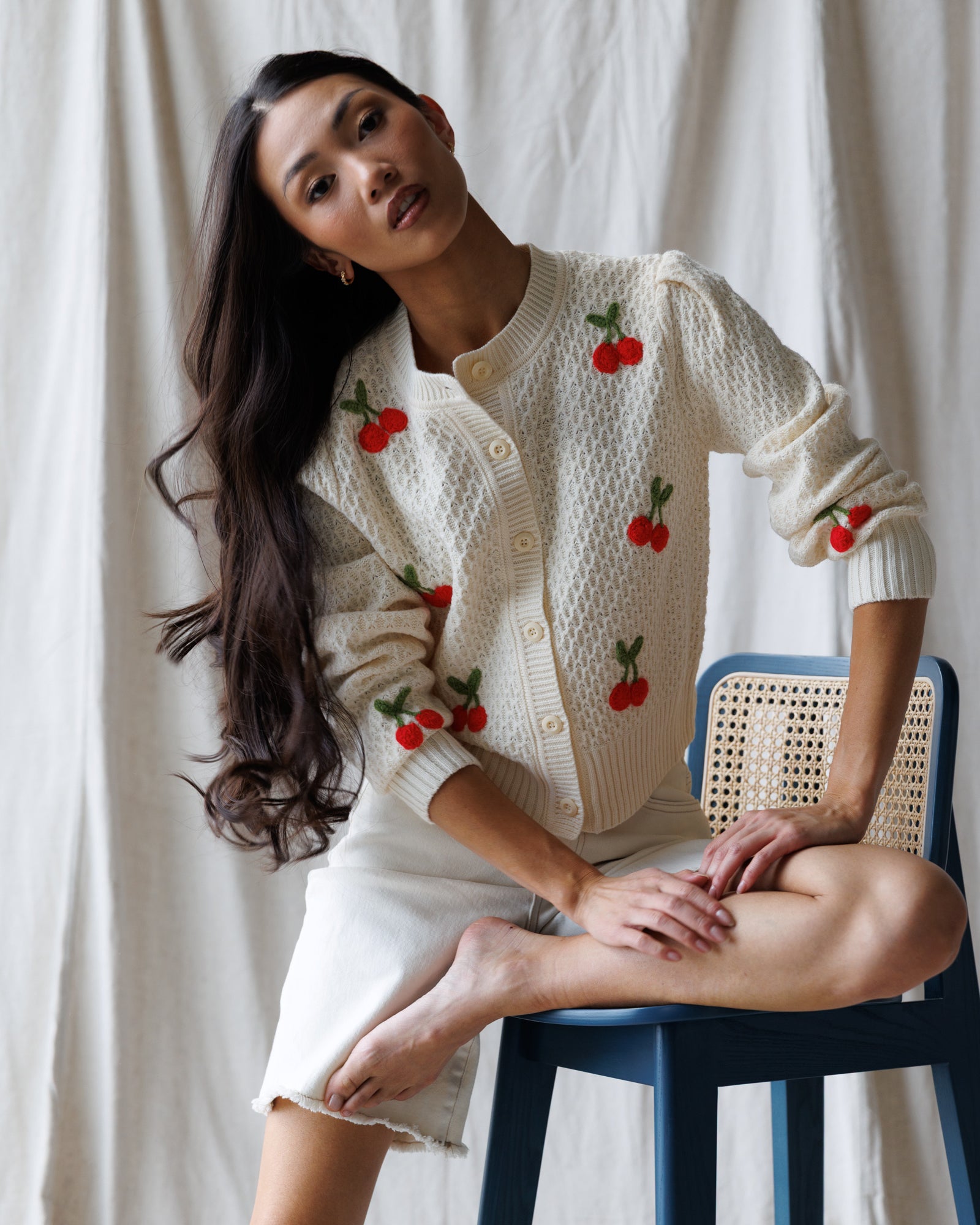 Woman wearing a white cardigan with red cherry patterns, sitting on a blue chair against a white curtain.