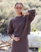 A woman wearing a textured brown knit mock-neck sweater and matching skirt stands outdoors in a dry, sandy environment with a rock and sunflowers in the background.