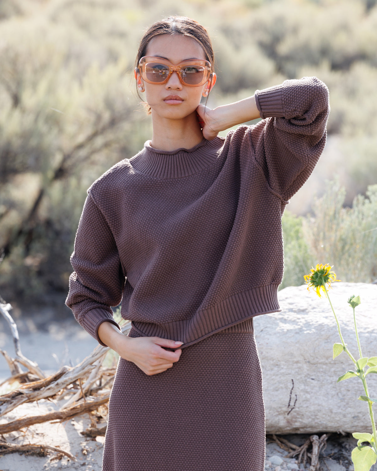 A woman wearing a textured brown knit mock-neck sweater and matching skirt stands outdoors in a dry, sandy environment with a rock and sunflowers in the background.