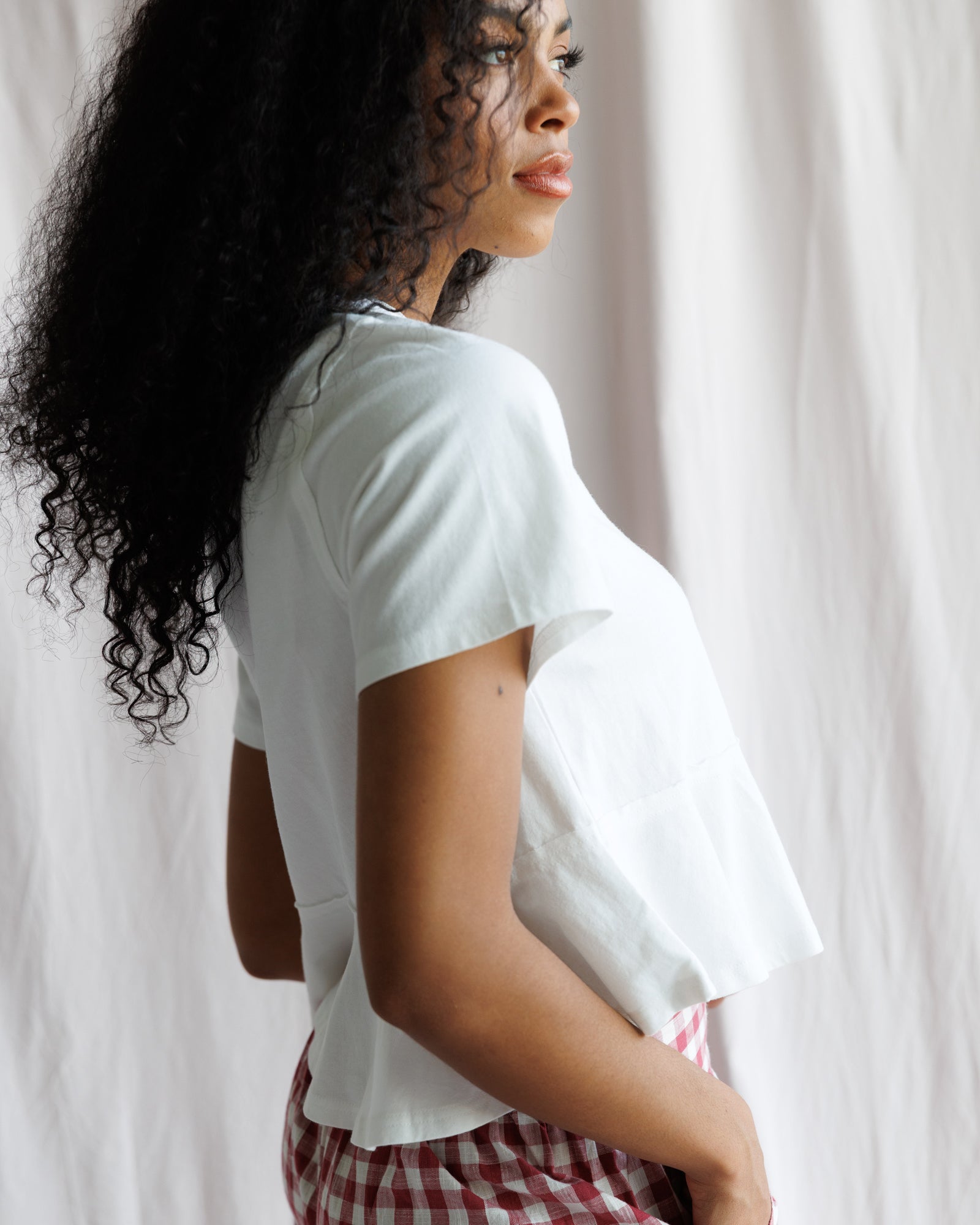 Woman wearing a white t-shirt and red checkered shorts standing against a white curtain.