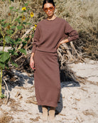 A woman stands outdoors in a sandy, brushy environment wearing a matching brown knit sweater and midi skirt set, along with beige ankle boots and orange-tinted sunglasses.