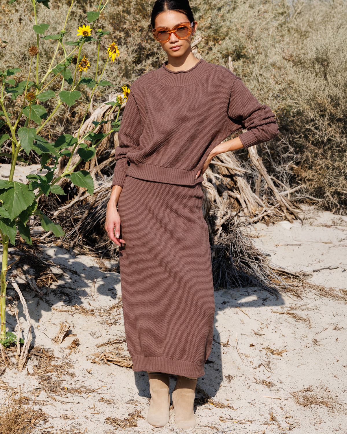 A woman stands outdoors in a sandy, brushy environment wearing a matching brown knit sweater and midi skirt set, along with beige ankle boots and orange-tinted sunglasses.