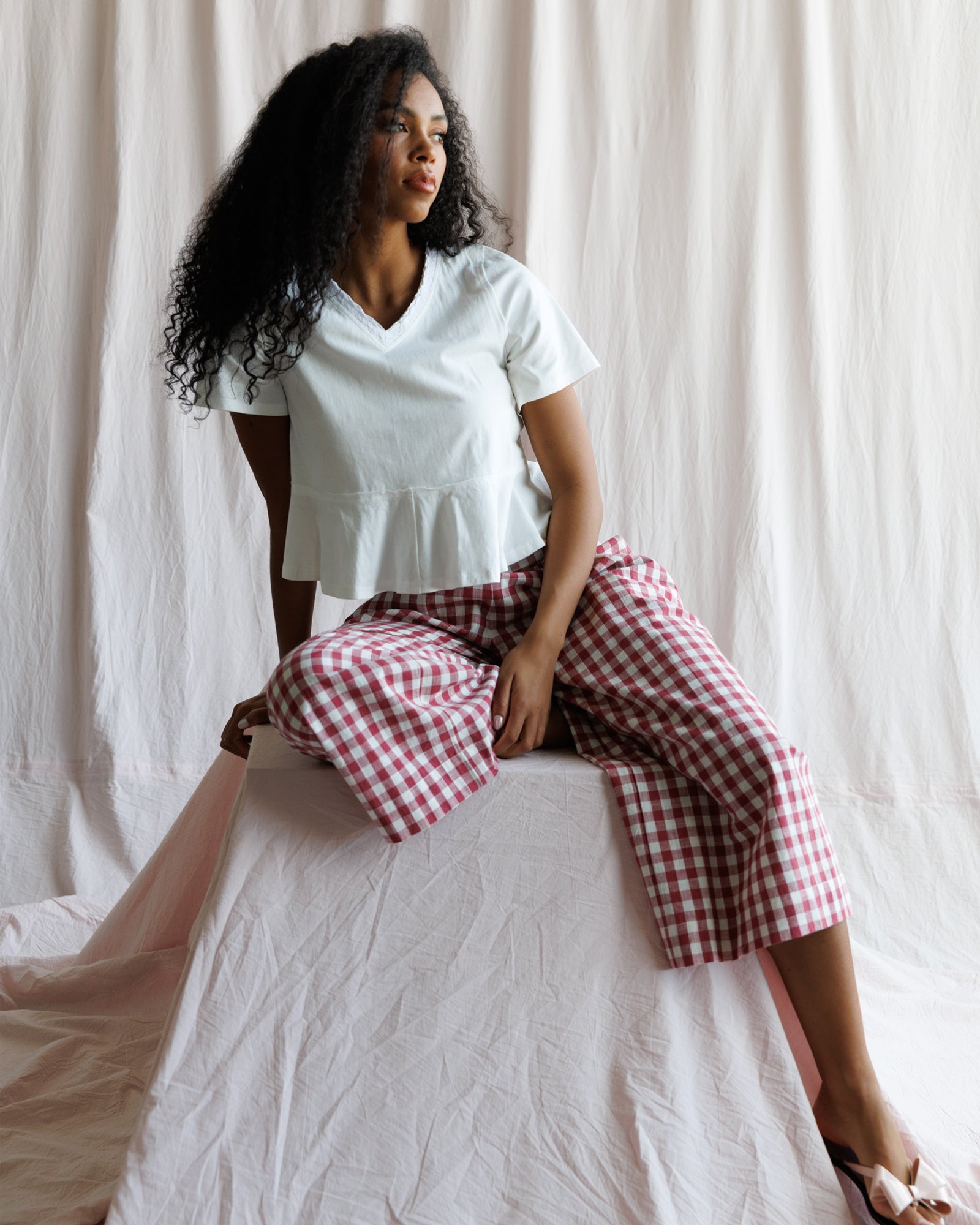 Woman wearing a white top and red checkered pants sitting on a white surface with a white curtain background.