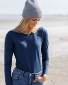 A woman wearing a navy blue long-sleeve crewneck shirt, blue jeans, and a light gray ribbed knit beanie, standing outdoors against a soft-focus sandy background.