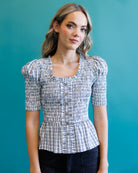 A woman wearing a short-sleeve button-front blouse with a blue, white, and gray plaid pattern and puff sleeves, posed against a solid teal background.