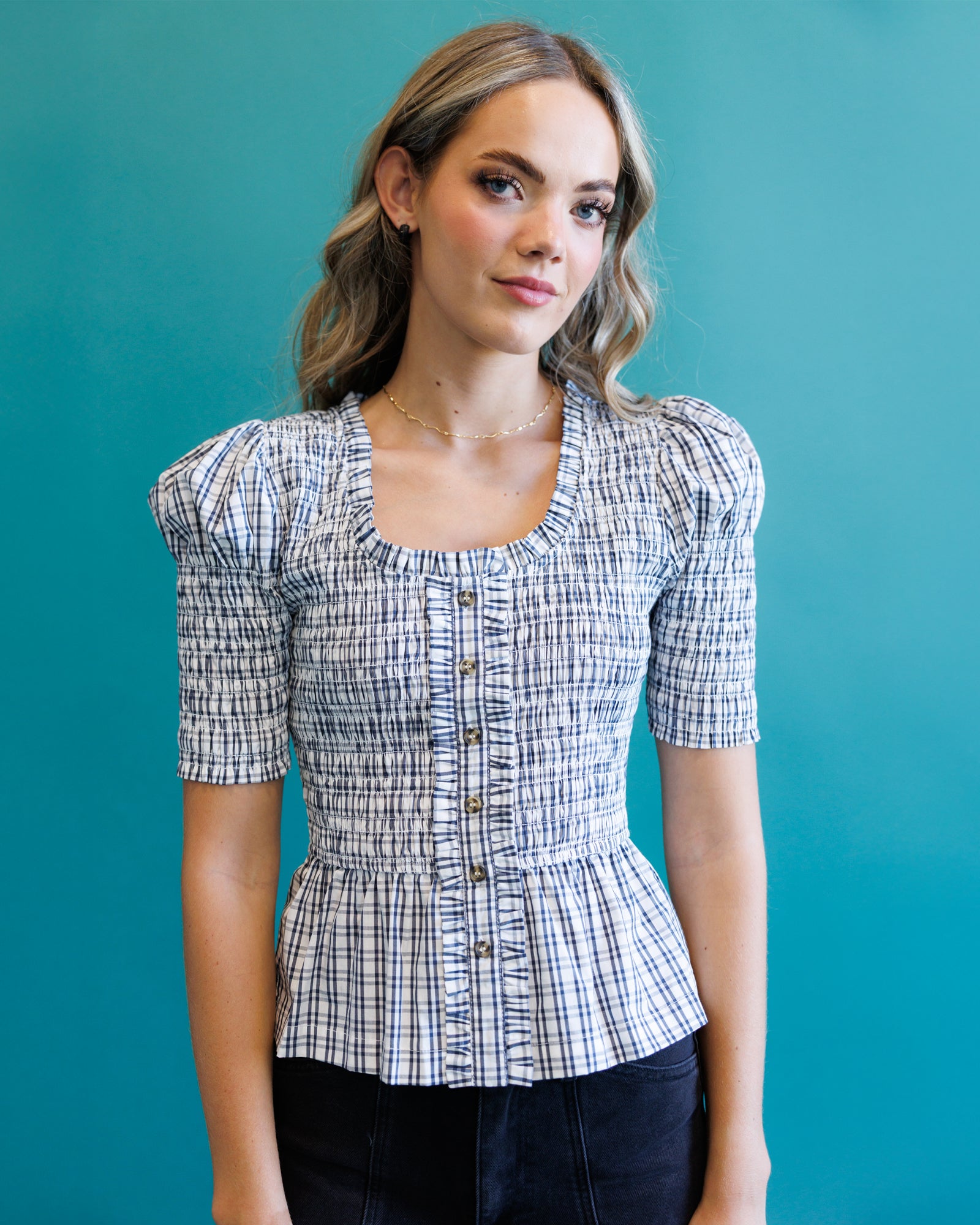 A woman wearing a short-sleeve button-front blouse with a blue, white, and gray plaid pattern and puff sleeves, posed against a solid teal background.