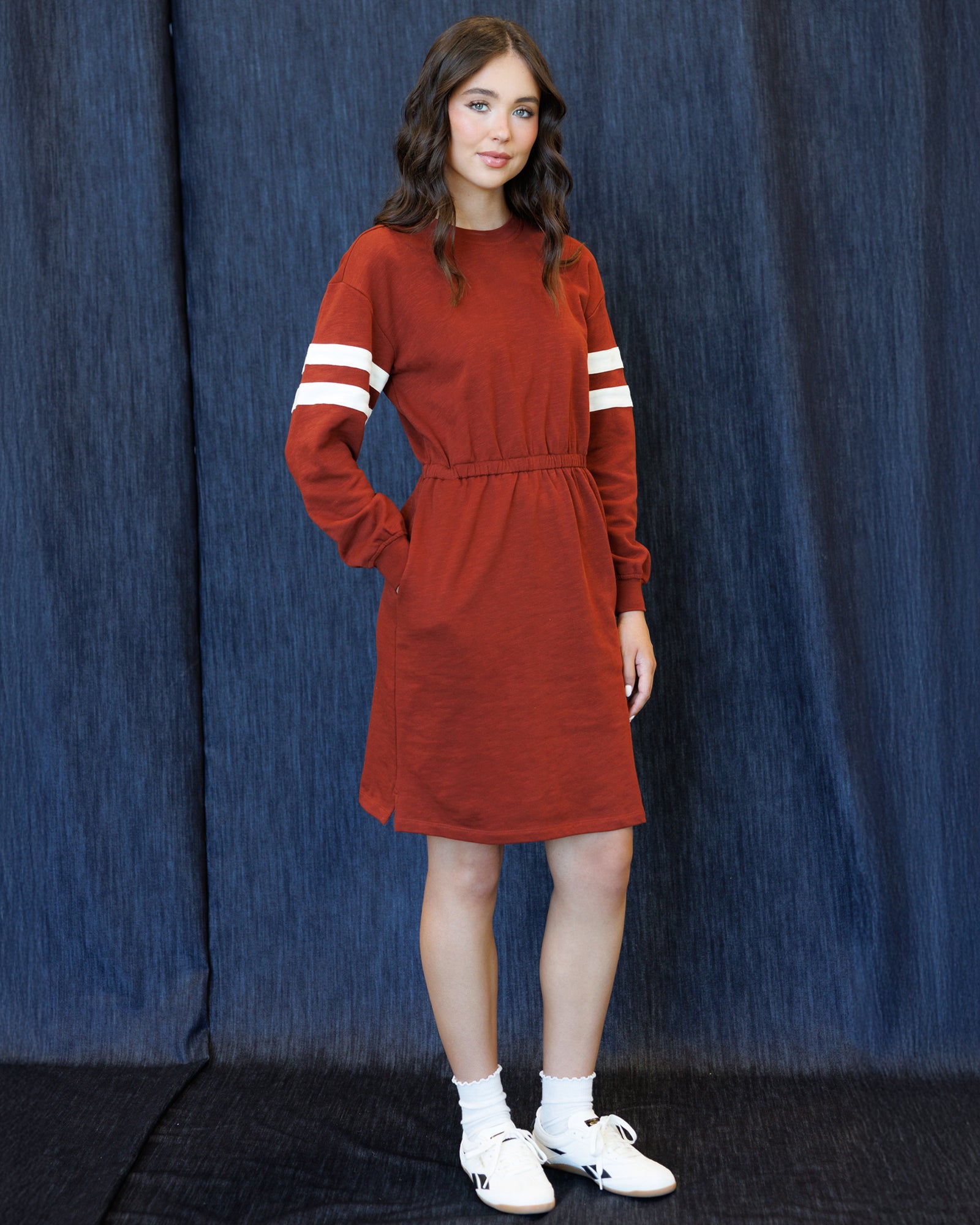 A young woman modeling a rust-red long-sleeve knit dress with white stripes on the sleeves, standing against a dark blue textured backdrop.