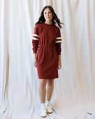 A woman modeling a rust-red long-sleeve knit dress with cream stripes on the sleeves, standing against a draped white fabric backdrop.