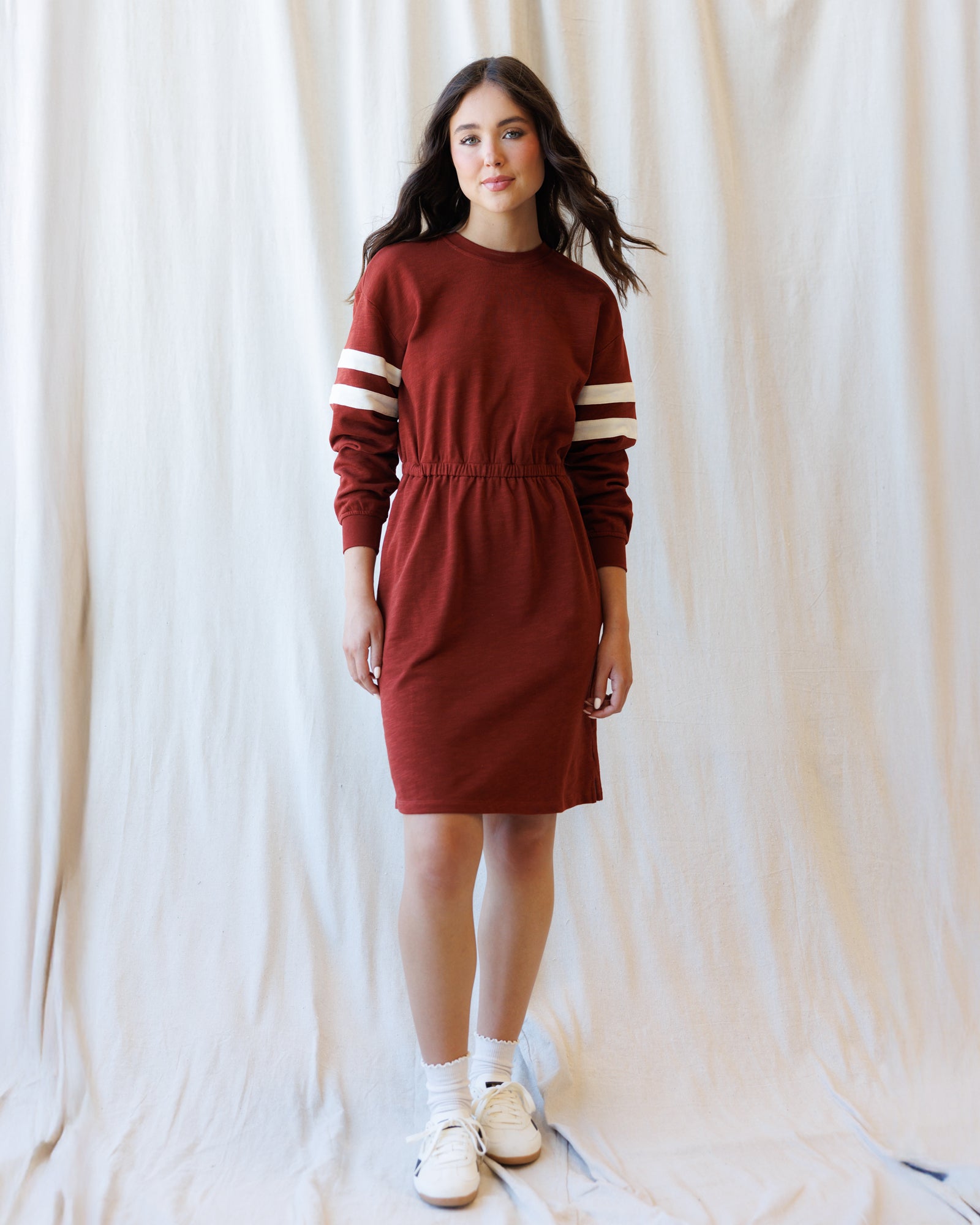 A woman modeling a rust-red long-sleeve knit dress with cream stripes on the sleeves, standing against a draped white fabric backdrop.
