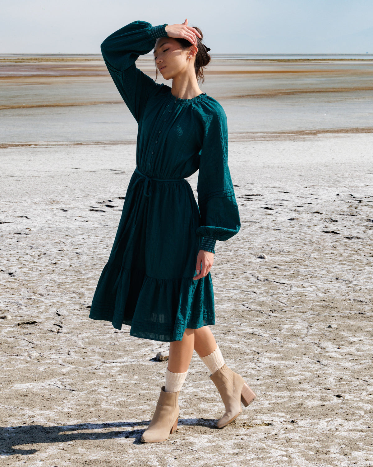 A woman stands on a dry, salt-flat landscape wearing a dark teal green long-sleeve midi dress with a tie waist and beige ankle boots.