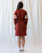 Back view of a woman modeling a rust-red long-sleeve dress with white stripes on the sleeves, standing against a draped white fabric backdrop.