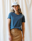 A woman wearing a faded blue crewneck t-shirt and a brown skirt, paired with a white baseball cap that has blue text, standing against a draped cream fabric backdrop.