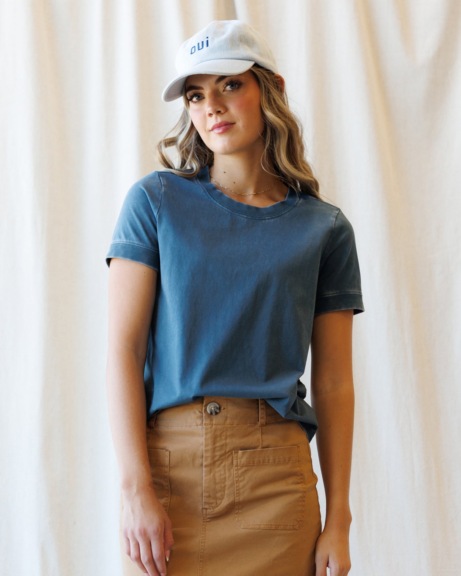 A woman wearing a faded blue crewneck t-shirt and a brown skirt, paired with a white baseball cap that has blue text, standing against a draped cream fabric backdrop.