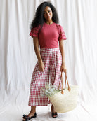 Woman wearing a red top and pink checkered pants holding a straw bag against a white background