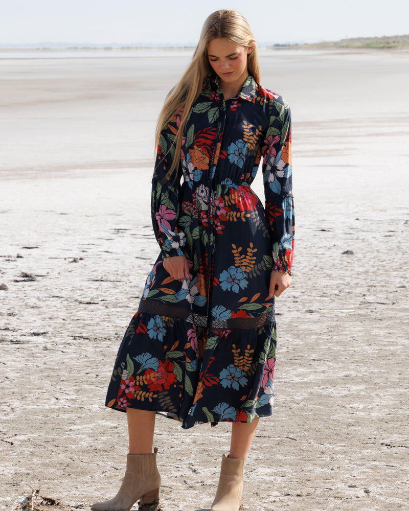 A woman stands on a sandy beach wearing a long-sleeve midi shirt dress with a colorful floral print on a dark navy background, paired with beige ankle boots.