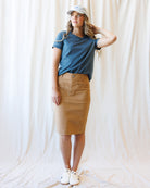 A woman modeling a blue short-sleeve t-shirt and a tan knee-length skirt, paired with a white baseball cap and white sneakers, standing against a draped white fabric backdrop.