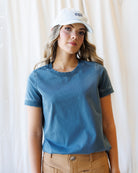 A woman wearing a washed slate blue short-sleeve crewneck t-shirt, tan pants, and a white baseball cap with the text 'oui' on it, standing in front of a soft white curtain background.