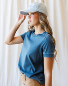 A woman modeling a blue short-sleeve t-shirt and a tan knee-length skirt, paired with a white baseball cap, standing against a draped white fabric backdrop.