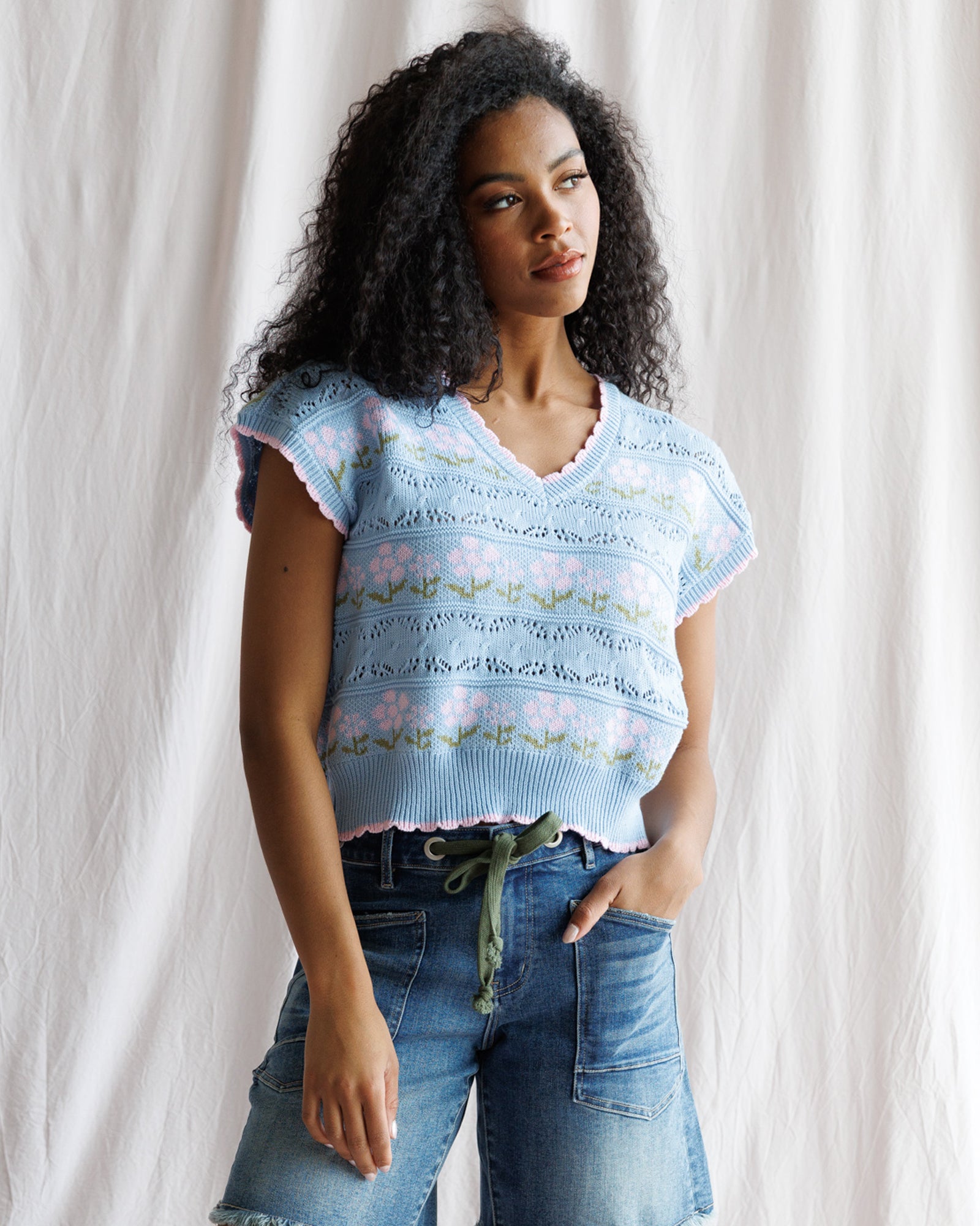 Woman wearing a light blue floral-patterned sleeveless top against a white curtain background