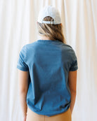 A woman modeling a blue short-sleeve t-shirt and a tan knee-length skirt, paired with a white baseball cap, standing against a draped white fabric backdrop.