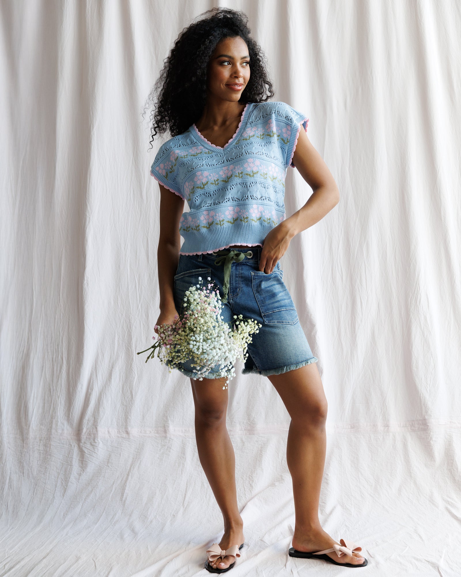 Woman wearing a light blue sleeveless top and denim shorts, holding flowers against a white background
