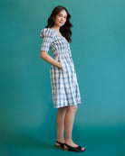 A woman modeling a short-sleeve blue and white plaid dress with a gathered waist, standing against a solid teal studio backdrop.