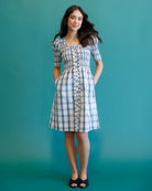 A woman modeling a short-sleeved blue and white plaid button-front dress with pockets, standing against a solid teal background.