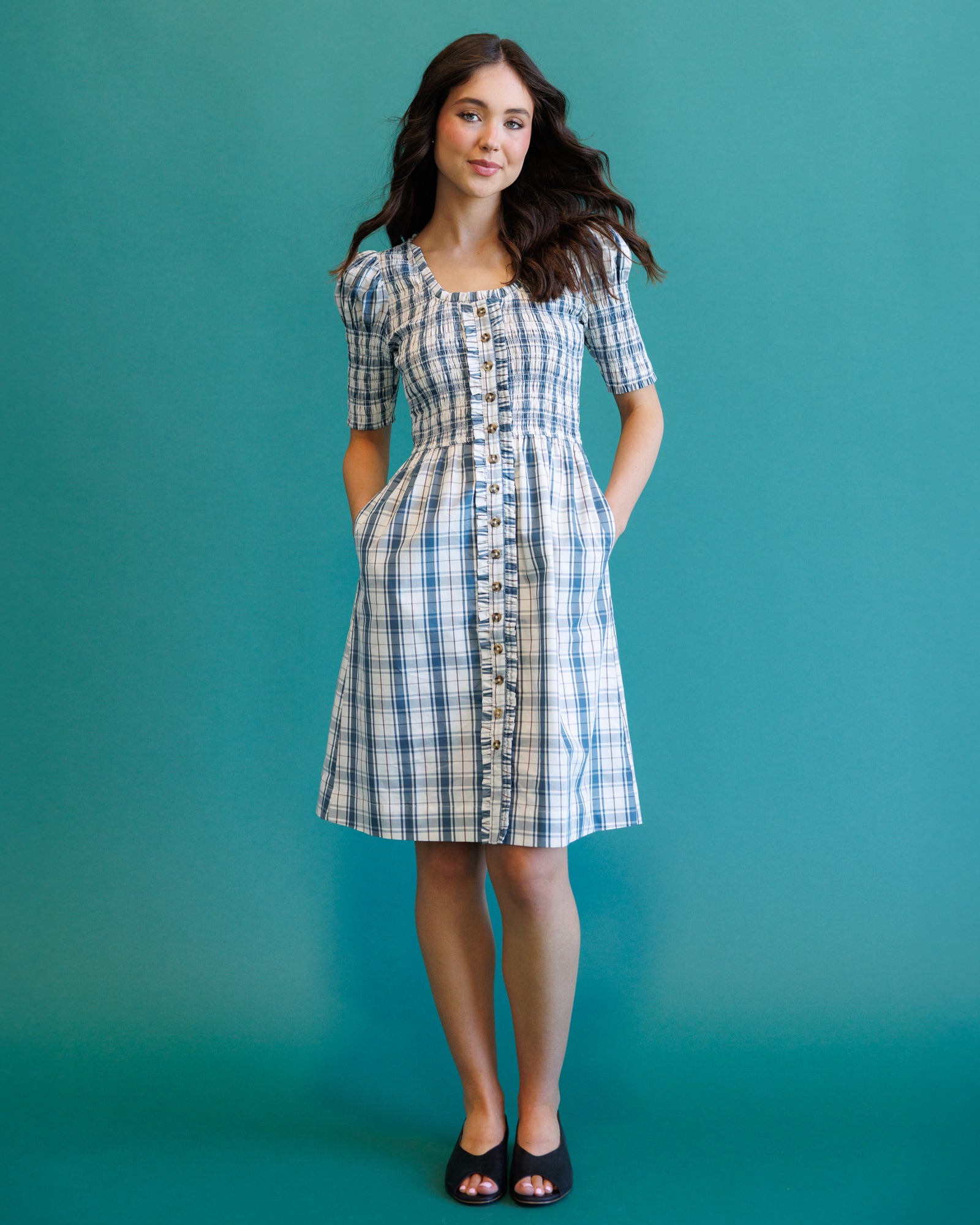 A woman modeling a short-sleeved blue and white plaid button-front dress with pockets, standing against a solid teal background.