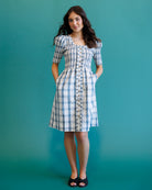 A woman modeling a short-sleeve plaid dress with a button-front bodice and side pockets, standing against a solid teal studio background.