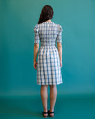 A woman viewed from behind wearing a short-sleeved blue and white plaid dress with puff sleeves and a gathered waist, standing against a solid teal background.