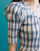 Close-up of a person wearing a smocked short-sleeve dress with a blue, white, and beige plaid pattern against a solid teal background.