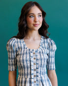 A woman wearing a blue, white, and beige plaid puff-sleeve dress with a buttoned neckline, posed against a solid teal background.