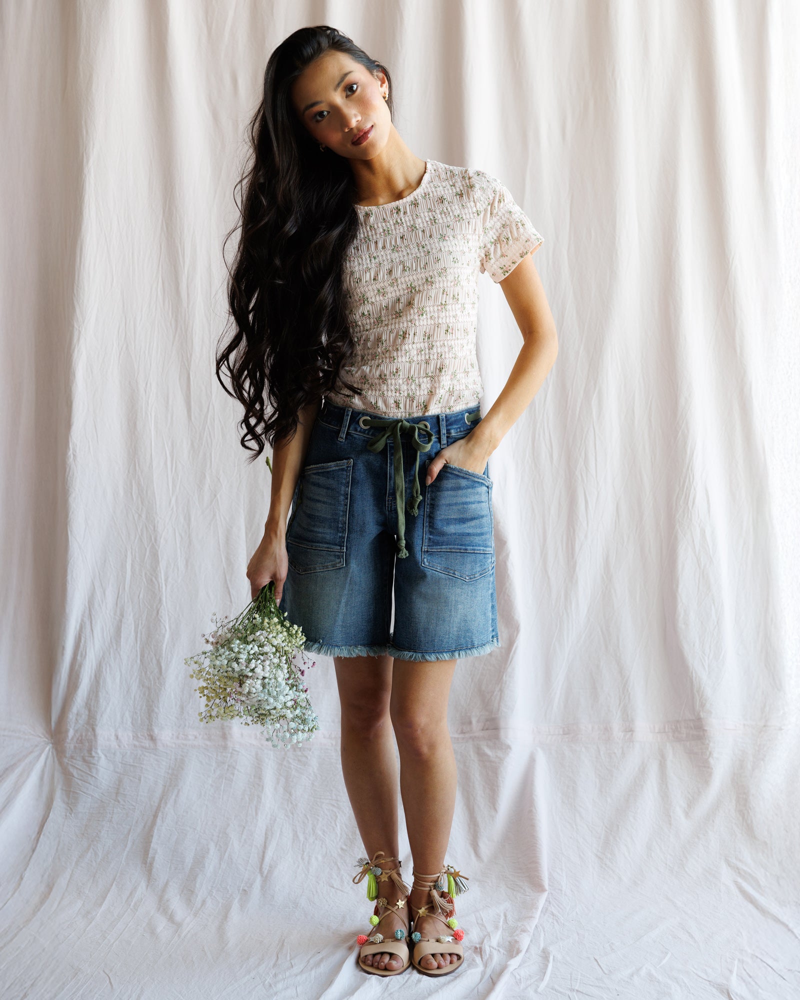 Woman wearing a floral top and denim skirt holding flowers against a white background
