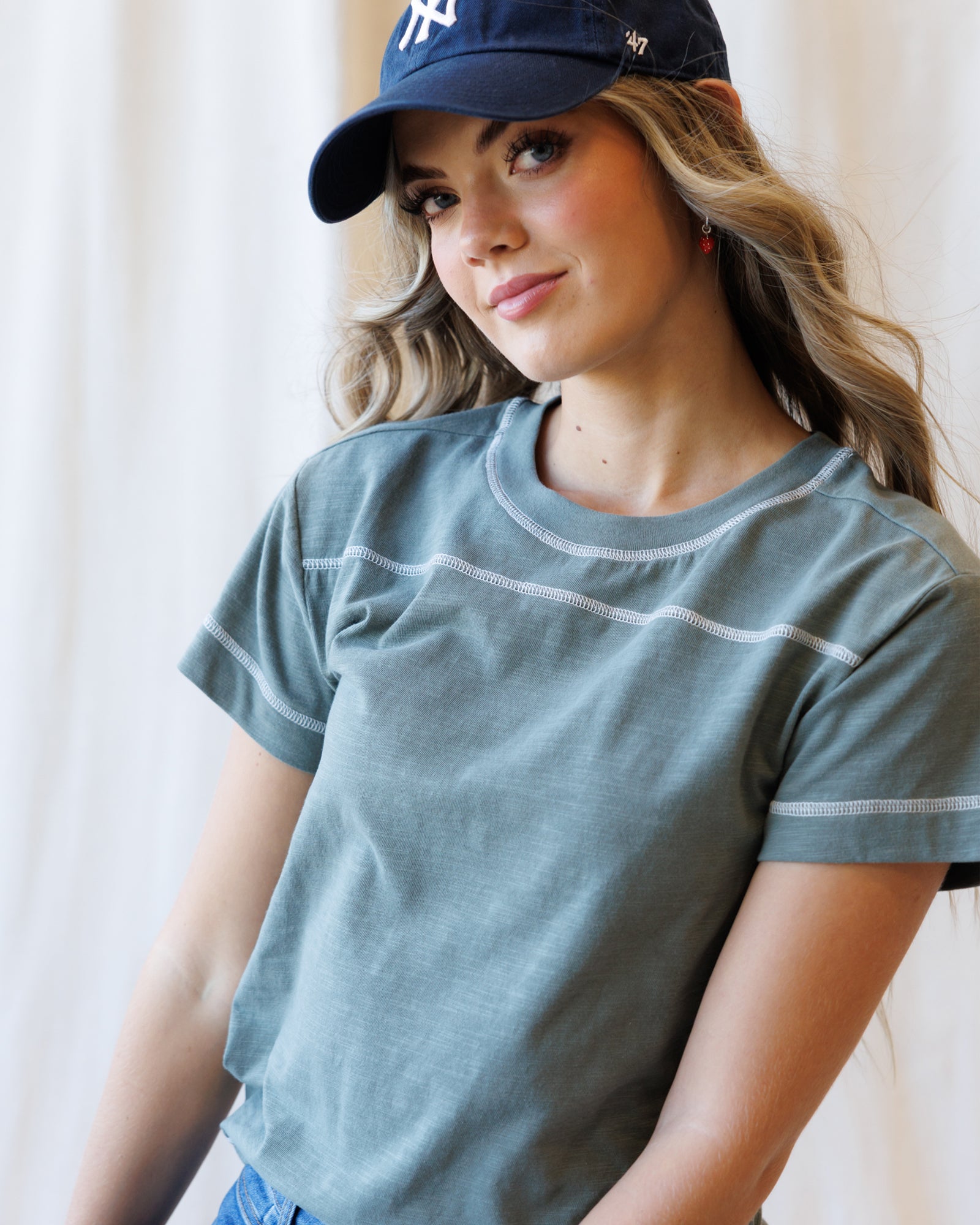 A woman wearing a slate blue-gray short-sleeve t-shirt with white contrast stitching and a navy baseball cap, photographed against a soft white curtain background.