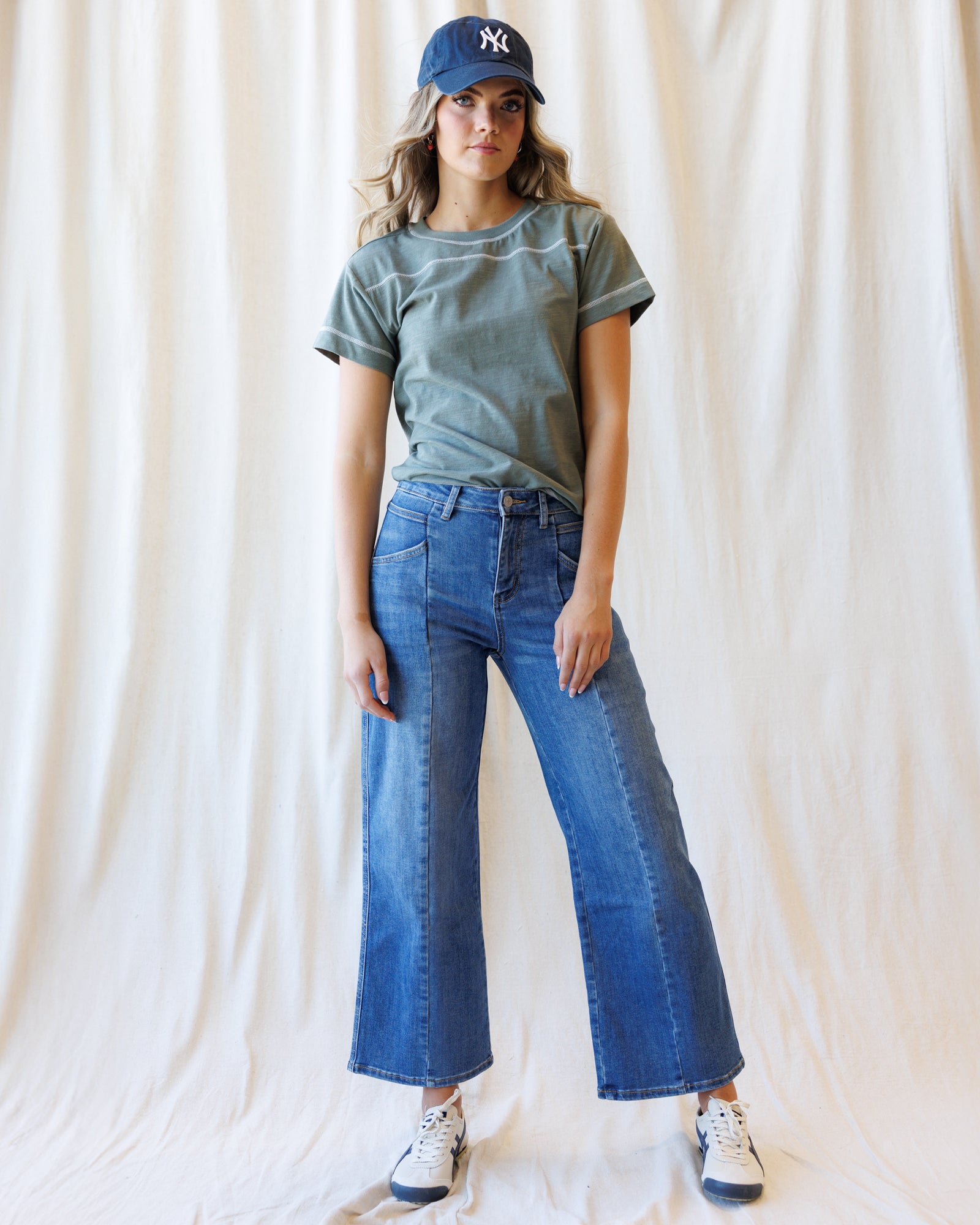 Woman wearing a green tshirt with blue jeans and a baseball cap standing in front of a canvas backdrop