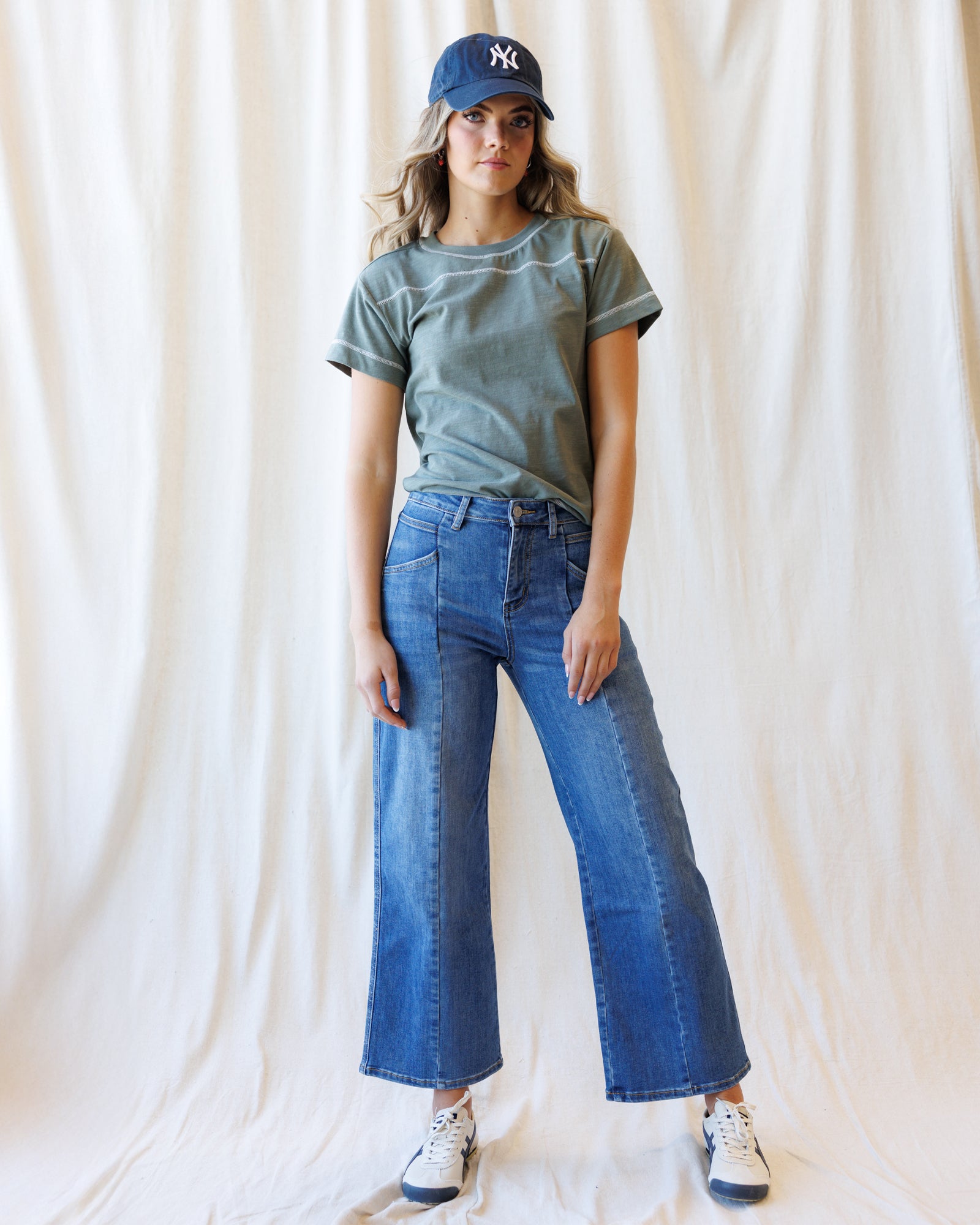 A woman stands against a draped cream fabric backdrop wearing a muted sage green short-sleeve t-shirt, wide-leg blue denim jeans, white sneakers, and a navy baseball cap with a white New York Yankees logo.