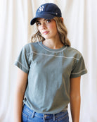 A woman wearing a faded sage green short-sleeve t-shirt with white contrast stitching, a navy blue New York Yankees baseball cap, and blue jeans, standing against a white curtain background.