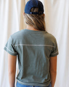 A person shown from the back wearing a muted sage green short-sleeve t-shirt with a thin white horizontal stripe across the upper back and a navy blue baseball cap, set against a soft white curtain background.