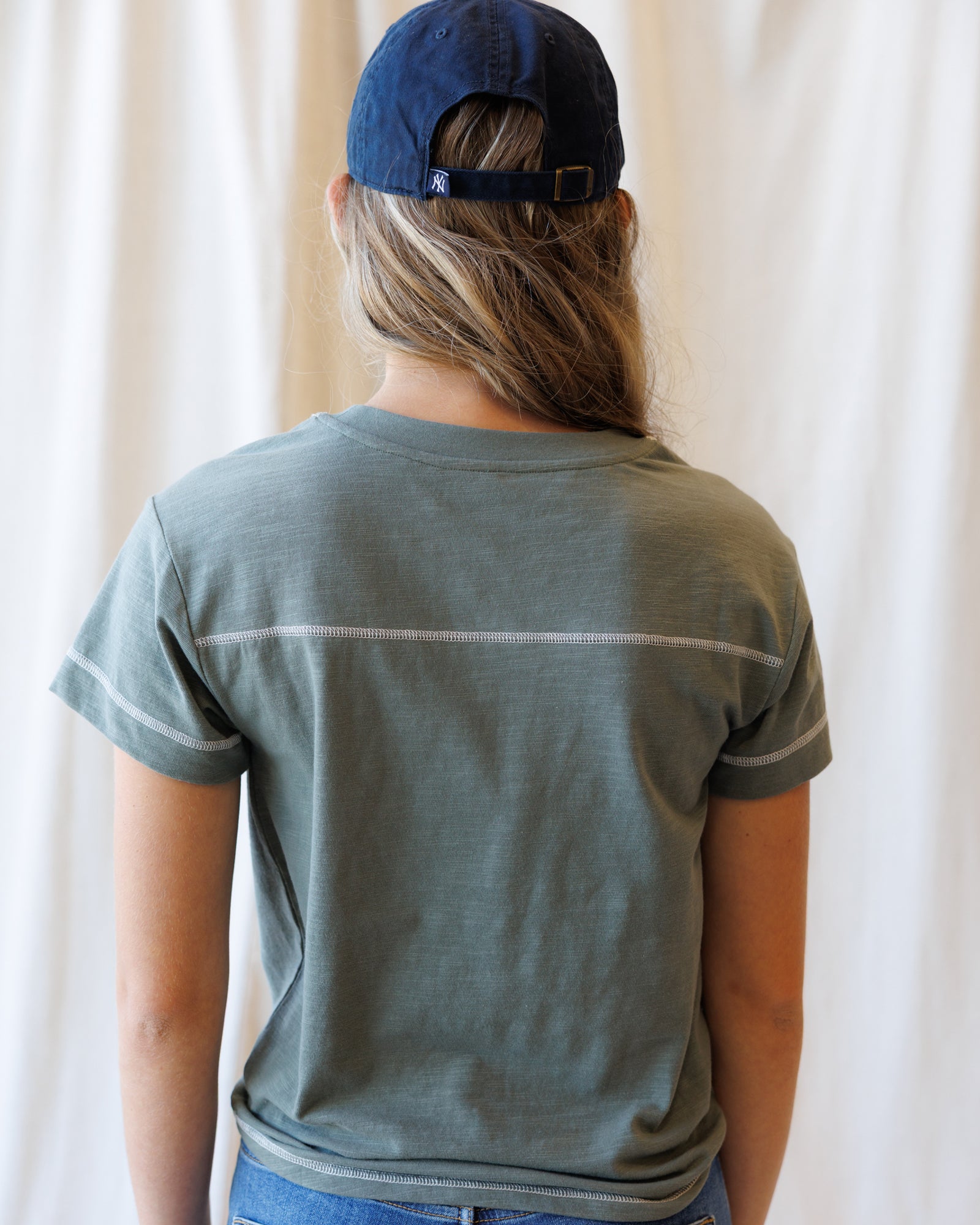 A person shown from the back wearing a muted sage green short-sleeve t-shirt with a thin white horizontal stripe across the upper back and a navy blue baseball cap, set against a soft white curtain background.