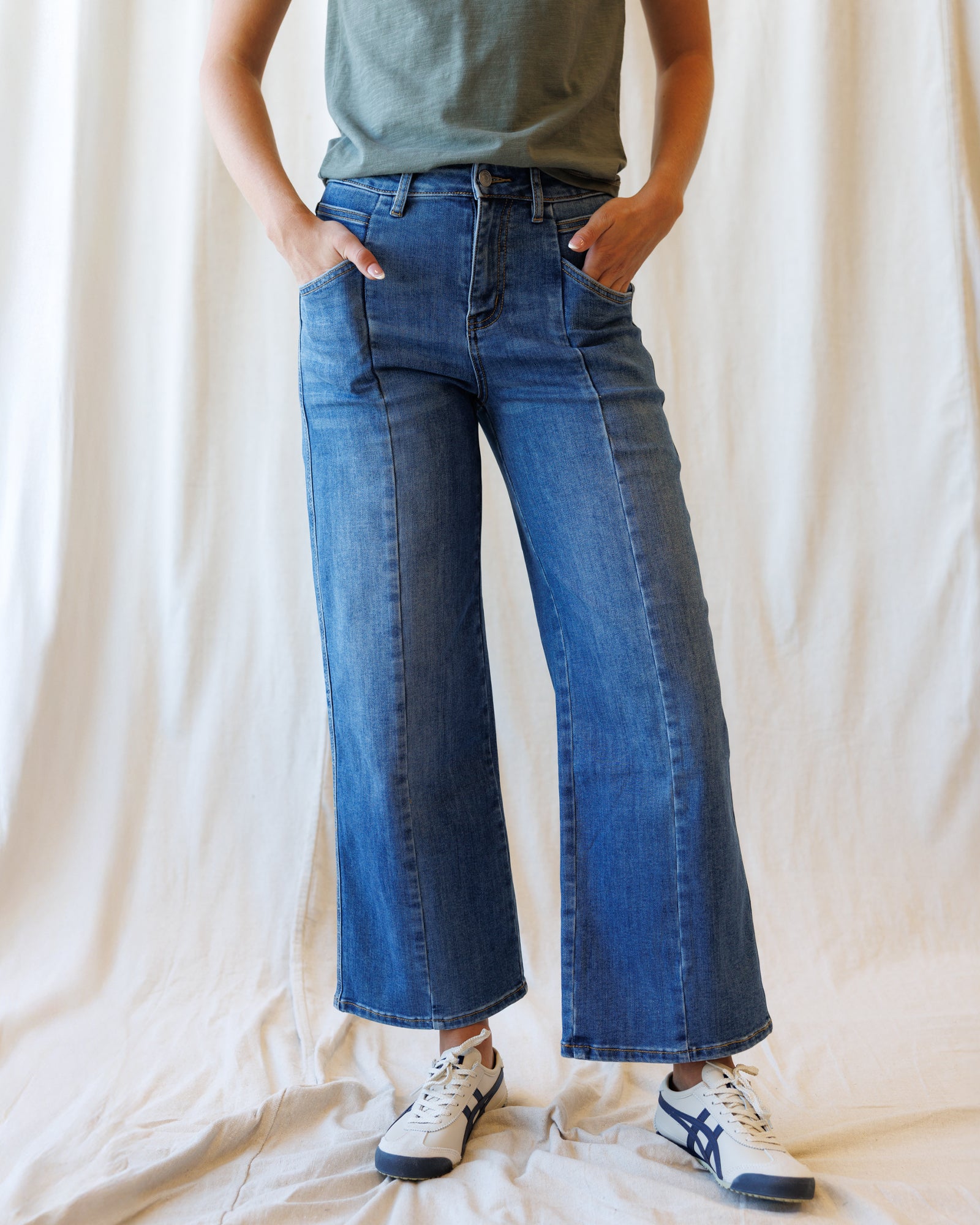 A person wearing blue wide-leg denim jeans with a muted green top and white sneakers, standing against a draped cream fabric backdrop.