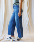 A person wearing high-waisted, wide-leg blue denim jeans with a faded wash, styled with a gray t-shirt and white sneakers with black stripes, standing against a draped cream fabric backdrop.