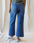 A person shown from the back wearing blue wide-leg cropped jeans, a gray top, and white sneakers with blue stripes, standing against a draped cream fabric backdrop.