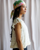 Woman wearing a colorful headscarf and white lace top against a plain background