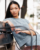 A woman wearing a light blue textured knit top and matching skirt, leaning on a dark metal bench in an outdoor setting.