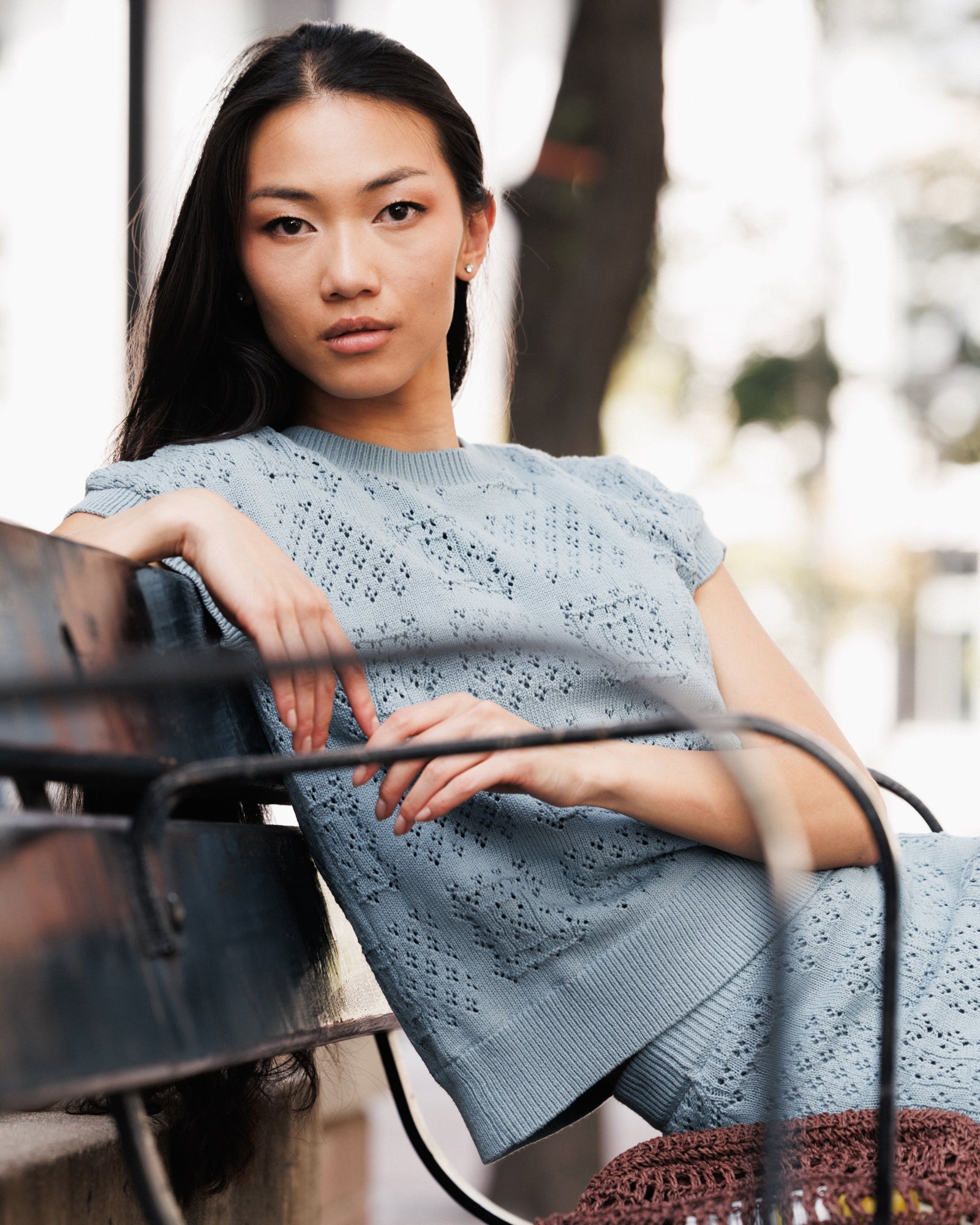 A woman wearing a light blue textured knit top and matching skirt, leaning on a dark metal bench in an outdoor setting.