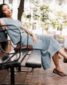 A woman wearing a light blue textured knit midi dress and matching top sits on a black metal park bench. She is wearing brown woven slip-on shoes and carrying a matching brown woven handbag.