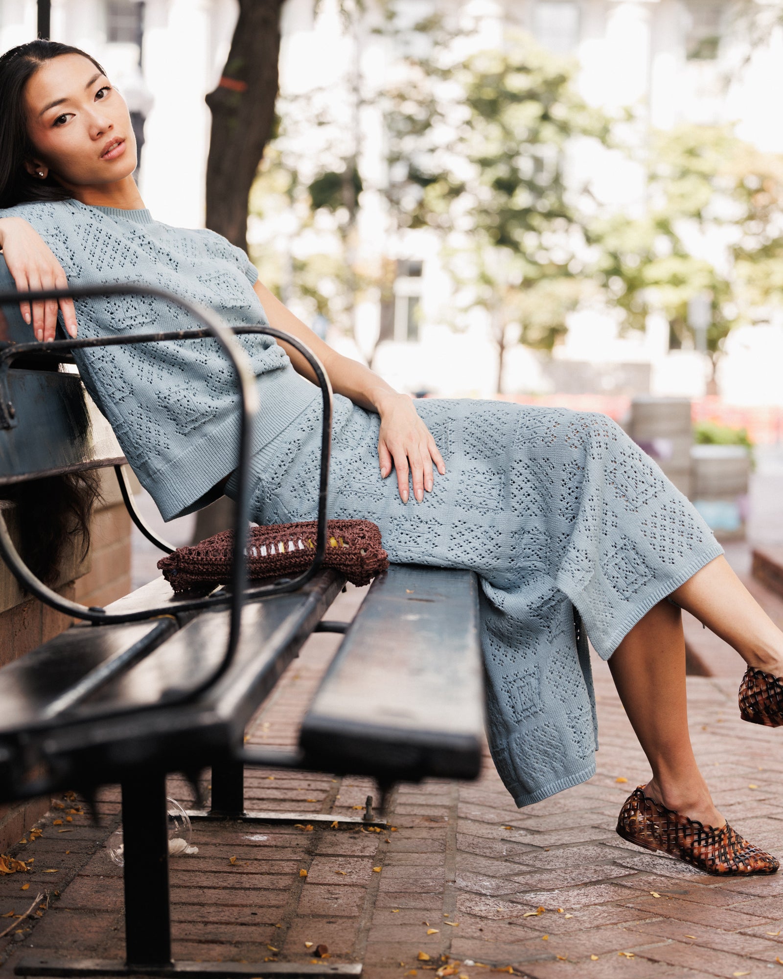 A woman wearing a light blue textured knit midi dress and matching top sits on a black metal park bench. She is wearing brown woven slip-on shoes and carrying a matching brown woven handbag.
