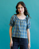 A woman modeling a blue and green checkered puff-sleeve blouse with a button-down front, paired with dark jeans, set against a solid teal background.